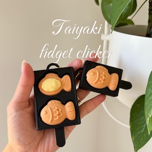 May include: Two black Taiyaki fidget clickers are held in a hand. One is open, revealing a yellow filling, and the other is closed. The fish-shaped toys are orange and have a black handle. The text "Taiyaki fidget clicker" is in the background.