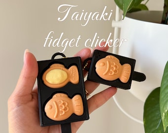 Peut inclure: Deux clickers anti-stress Taiyaki noirs sont tenus dans une main. L'un est ouvert, révélant une garniture jaune, et l'autre est fermé. Les jouets en forme de poisson sont orange et ont une poignée noire. Le texte "Taiyaki fidget clicker" est en arrière-plan.