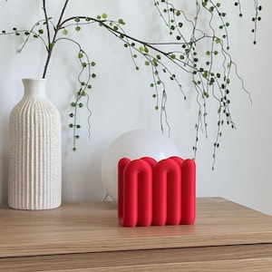 May include: A bright red, matte-finished sculptural object featuring a series of interconnected, rounded vertical tubes arranged in a continuous, wave-like pattern. The piece has a smooth, uniform texture and a bold, solid colour throughout its structure.