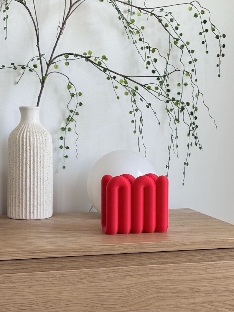 May include: A bright red, matte-finished sculptural object featuring a series of interconnected, rounded vertical tubes arranged in a continuous, wave-like pattern. The piece has a smooth, uniform texture and a bold, solid colour throughout its structure.