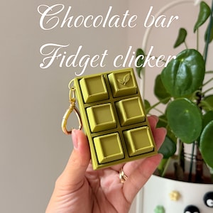 May include: A rectangular olive green fidget clicker shaped like a chocolate bar with six square buttons, one featuring a small leaf icon. The device includes a gold-coloured metal keychain attachment. The text "Chocolate bar Fidget clicker" is visible.