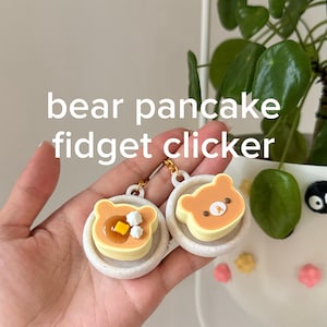 Könnte beinhalten: Zwei Bären-Pfannkuchen-Fidget-Clicker mit hellbraunen, bärenförmigen Pfannkuchen auf weiß gesprenkelten Tellern. Ein Pfannkuchen ist mit einem gelben Butterstück und weißen Tupfen belegt, der andere zeigt ein Bärengesicht. Text: bear pancake fidget clicker.
