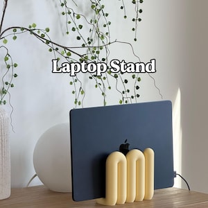 May include: A pale yellow, wavy, U-shaped laptop stand holding a dark grey laptop. The stand features a series of rounded, vertical, connected loops that support the device in an upright position. The text "Laptop Stand" is visible above.