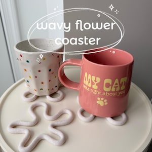 May include: A pink ceramic mug with the text "MY CAT was right about you" and a paw print, resting on a white coaster. A second mug with colorful star and flower designs sits on a matching coaster. A wavy flower coaster is also present.