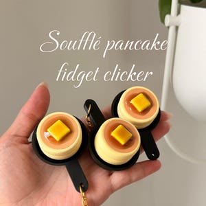 May include: Three Soufflé pancake fidget clickers with a yellow square of butter on top, in a black pan, attached to a gold keychain. The text "Soufflé pancake fidget clicker" is in white script.