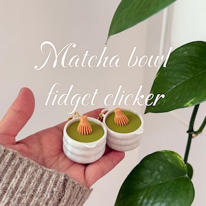 May include: Two white ceramic Matcha bowl fidget clickers with a green interior and a small orange whisk. The image includes the text "Matcha bowl fidget clicker". The clickers are held in a hand, with a green plant in the background.