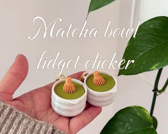 May include: Two white ceramic Matcha bowl fidget clickers with a green interior and a small orange whisk. The text "Matcha bowl fidget clicker" is in the image. The clickers are held in a hand, with a green plant in the background.