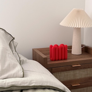 May include: A bright red, U-shaped, wavy decorative object with a matte finish, positioned next to a beige pleated lamp with a textured base on a wooden nightstand with two drawers featuring woven panels and thin metal handles.