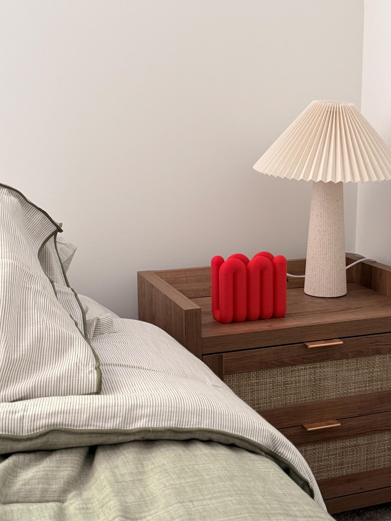 May include: A bright red, U-shaped, wavy decorative object with a matte finish, positioned next to a beige pleated lamp with a textured base on a wooden nightstand with two drawers featuring woven panels and thin metal handles.