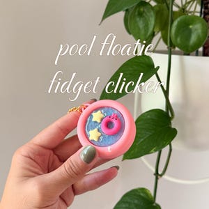 May include: A pink fidget clicker with a pool floatie design. The clicker features a blue interior with yellow stars and a pink donut-shaped floatie. The text "pool floatie fidget clicker" is written in cursive.