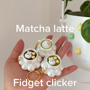 May include: Three white cup-shaped fidget clickers with gold keychains. Two cups contain green matcha latte with white whipped cream, a small white rabbit figure, and yellow stars. One cup contains dark brown liquid with a white rabbit and stars.