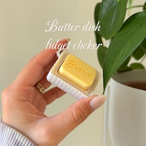 May include: A yellow butter-shaped fidget clicker with the word "Butter" printed on it, held in a white rectangular dish. The text "Butter dish fidget clicker" is written in cursive. The background includes green leaves and a white pot.