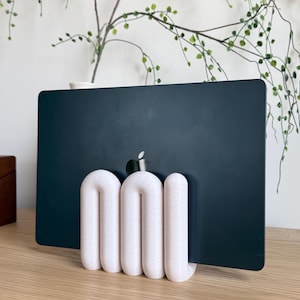 May include: A dark grey laptop with a silver Apple logo is held upright by a white, textured, U-shaped stand. The stand features three rounded, interconnected vertical loops that provide a stable base for the device to rest in a vertical position.