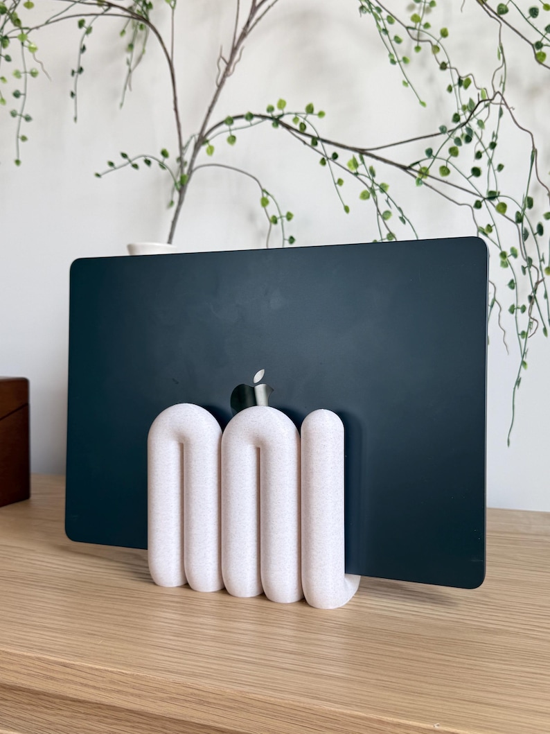 May include: A dark grey laptop with a silver Apple logo is held upright by a white, textured, U-shaped stand. The stand features three rounded, interconnected vertical loops that provide a stable base for the device to rest in a vertical position.