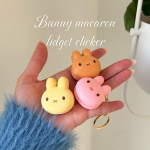May include: Three bunny-shaped fidget clickers in yellow, orange, and pink, held in a hand. Each has a different color and a small gold keyring attached to the pink one. The text "Bunny macaron fidget clicker" is in the background.