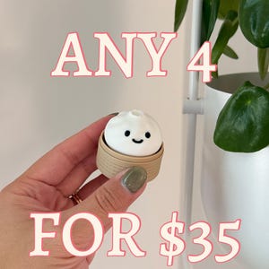May include: A white dumpling with a smiling face sits in a small, tan bamboo steamer. The text "ANY 4 FOR $35" is displayed in pink letters with a white outline. A hand holds the dumpling, with a plant in the background.