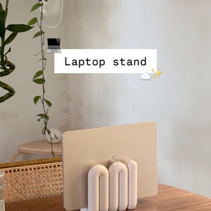 May include: A matte off-white laptop stand with a series of rounded, vertical, tube-like supports holding a gold-colored laptop in a vertical position. The stand features the text Laptop stand with a small laptop icon, a cloud, and stars.