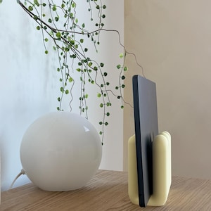 May include: A pale yellow, rounded, multi-slotted stand holding a thin, dark grey electronic tablet in an upright position. The stand features a smooth, matte finish with vertical grooves designed to support the device securely in a vertical orientation.
