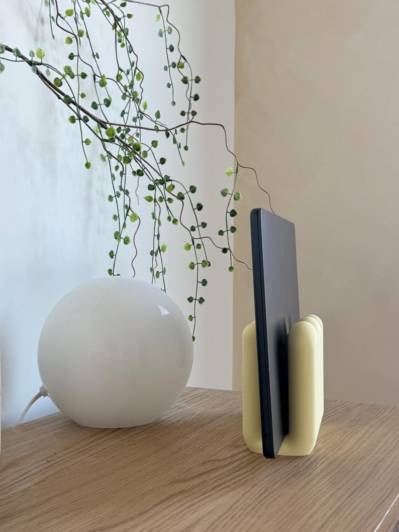 May include: A pale yellow, rounded, multi-slotted stand holding a thin, dark grey electronic tablet in an upright position. The stand features a smooth, matte finish with vertical grooves designed to support the device securely in a vertical orientation.