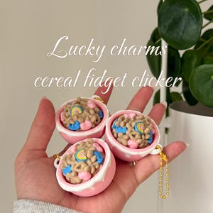 May include: Three pink and white Lucky Charms cereal fidget clickers. Each charm is a small bowl filled with cereal pieces in various colors. The charms have gold-colored chain attachments and the text "Lucky charms cereal fidget clicker".