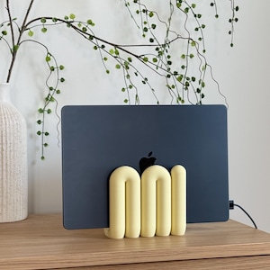 May include: A pale yellow, tubular, U-shaped laptop stand holding a dark grey laptop. The stand features a series of connected, rounded, arch-like forms that provide a stable base for the device, which displays a black Apple logo on its surface.