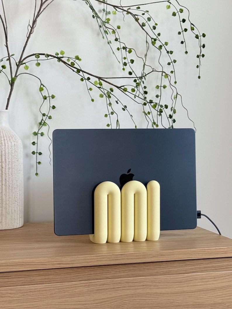 May include: A pale yellow, tubular, U-shaped laptop stand holding a dark grey laptop. The stand features a series of connected, rounded, arch-like forms that provide a stable base for the device, which displays a black Apple logo on its surface.