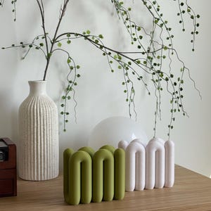 May include: Two sculptural objects with a repeating arched, tubular design. One object is a solid olive green colour with a smooth finish, while the other is a textured, off-white colour. Both items feature a series of connected, rounded loops.