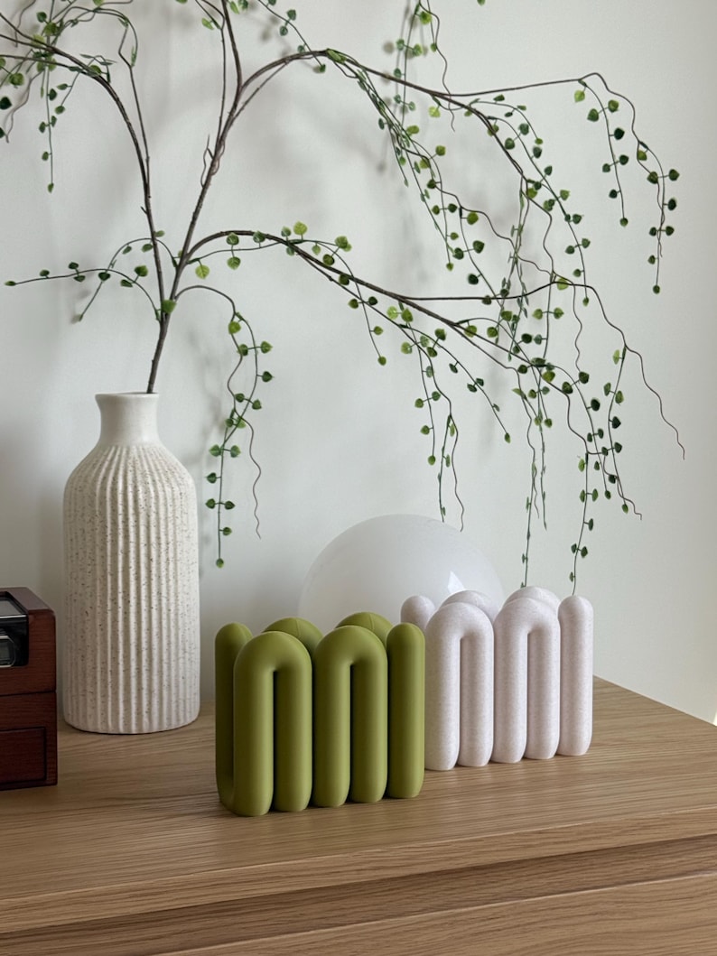 May include: Two sculptural objects with a repeating arched, tubular design. One object is a solid olive green colour with a smooth finish, while the other is a textured, off-white colour. Both items feature a series of connected, rounded loops.