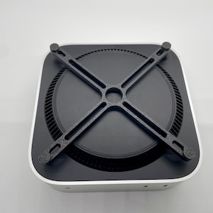 Könnte beinhalten: Ein schwarzer Metallständer mit vier Armen in X-Form, der für die Unterstützung eines Mac mini-Computers konzipiert ist. Der Ständer ist an der Unterseite des Mac mini befestigt, einem kleinen, quadratischen Computer mit schwarzer Oberseite und silberner Unterseite.