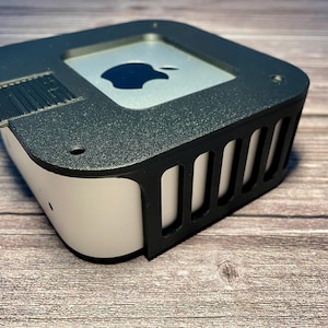 Könnte beinhalten: Ein schwarzes Plastikgehäuse mit einem Lüftungsdesign für einen kleinen, weißen Computer mit einem Apple-Logo auf der Oberseite.