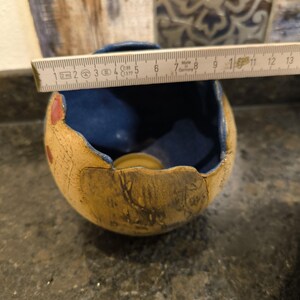 以下が含まれることがあります： 卵の形をした小さなセラミック製のキャンドルホルダー。ホルダーはひびが入っており、外側は青い釉薬、内側は黄色の釉薬で覆われています。ホルダーの直径は約8cmです。