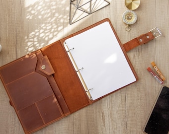 May include: A brown leather binder featuring a three-ring metal mechanism, a snap-button pocket, and a strap closure. The interior includes multiple slots for organization, holding blank white paper sheets within the metal rings.