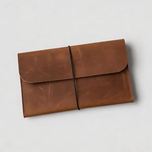May include: A brown leather portfolio with a flap closure and a black elastic band. The rectangular case is made of textured leather and is designed to hold documents or a tablet.