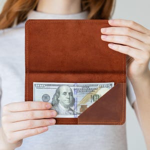 Op de afbeelding: Een bruine lederen portemonnee met een rechthoekig vak met een diagonale opening. Een biljet van honderd dollar zit in het vak en toont het portret van Benjamin Franklin en de tekst 100 United States of America.