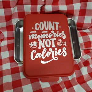 Puede incluir: Una bandeja para hornear rectangular roja con interior plateado y tapa roja a juego. La tapa presenta el texto blanco "COUNT memories NOT calories" en una fuente decorativa. La bandeja está sobre un mantel a cuadros rojos y blancos.