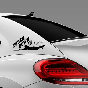 K&ouml;nnte beinhalten: Ein wei&szlig;es Auto mit einem schwarzen Vinyl-Aufkleber auf der Heckscheibe. Der Aufkleber lautet "Touch Down!!!" und zeigt eine Person, die ins Wasser springt.