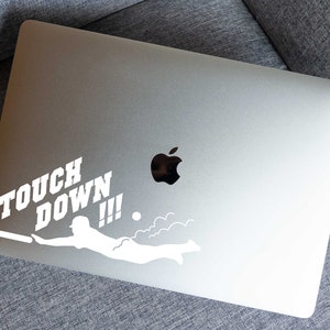 K&ouml;nnte beinhalten: Ein wei&szlig;er Vinyl-Aufkleber mit einer Silhouette einer Person, die mit einem Fu&szlig;ball taucht, und den Worten "TOUCH DOWN!!!" auf einem silbernen Laptop.