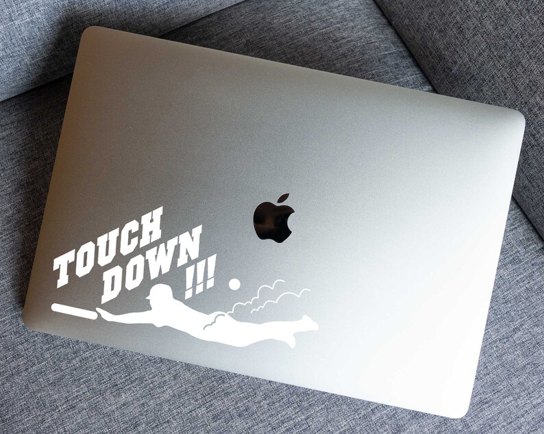K&ouml;nnte beinhalten: Ein wei&szlig;er Vinyl-Aufkleber mit einer Silhouette einer Person, die mit einem Fu&szlig;ball taucht, und den Worten "TOUCH DOWN!!!" auf einem silbernen Laptop.