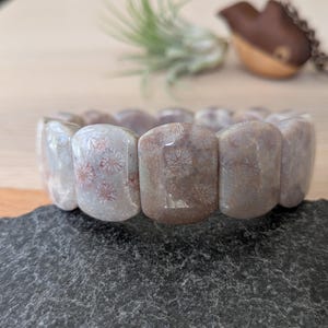 May include: A stretch bracelet made of polished, rectangular coral stones. The stones are primarily gray with subtle pink and white patterns. The bracelet is displayed on a dark gray, textured surface, with a wooden background.