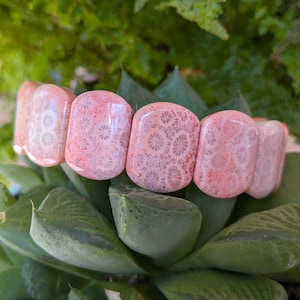 May include: A coral-colored bracelet with a stretch design. The bracelet features large, rectangular beads with a detailed, patterned surface. The beads are a mix of pink and white, and the bracelet is set against a backdrop of green foliage.