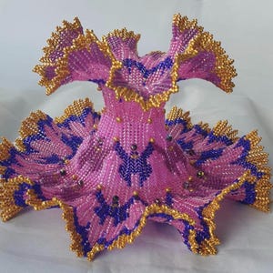 Może przedstawiać: Ręcznie wykonana, dekoracyjna rzeźba w kształcie kwiatu. Rzeźba wykonana jest z różowych, złotych i niebieskich koralików. Krawędzie są obszyte złotymi koralikami. Rzeźba ma unikalny, teksturowany wygląd.