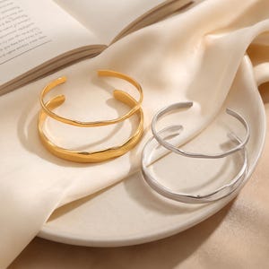 Puede incluir: Dos pulseras de puño abiertas. Una es dorada, la otra plateada. La pulsera dorada tiene un aspecto texturizado y martillado. La pulsera plateada tiene un acabado liso. Ambas pulseras se exhiben en un plato blanco.