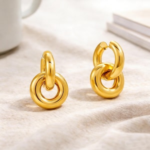 May include: A pair of gold-toned hoop earrings, each featuring a polished, thick circular band connected to a smaller, matching circular ring. The earrings have a smooth, reflective metallic finish and a classic, interlocking design with a hinged closure.