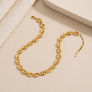 May include: A gold-toned necklace featuring a series of repeating cowrie shell-shaped links connected in a continuous chain. The necklace includes a short extender chain with a small metal bar at the end, providing an adjustable fit for the wearer.