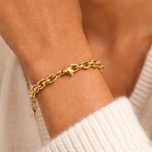 May include: A gold-colored chain bracelet with a double-strand design. The bracelet features oval-shaped links and a lobster clasp closure. The jewelry is worn on a wrist, showcasing its polished finish and elegant style.