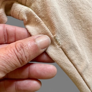 May include: A light beige cotton fabric garment featuring a visible seam construction with reinforced stitching along the edge. The material has a textured, woven appearance with a consistent neutral tone throughout the visible section of the textile.