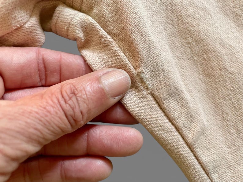 May include: A light beige cotton fabric garment featuring a visible seam construction with reinforced stitching along the edge. The material has a textured, woven appearance with a consistent neutral tone throughout the visible section of the textile.