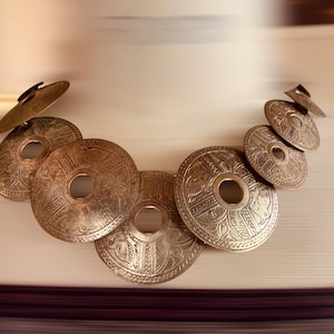 Pode incluir: Uma coleção de discos decorativos antigos de latão com intrincados padrões em relevo. Os discos variam em tamanho e estão dispostos em um design curvo em cascata. Os tons metálicos quentes criam uma estética vintage.