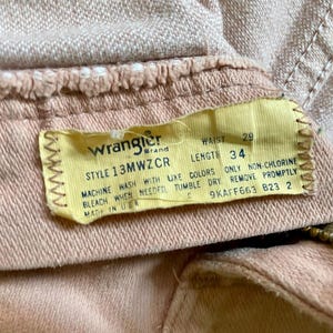May include: A pair of light tan denim pants featuring a yellow interior tag with the Wrangler brand logo, style number 13MWZCR, and printed measurements of 29 inch waist and 34 inch length. The tag includes care instructions and Made in USA text.
