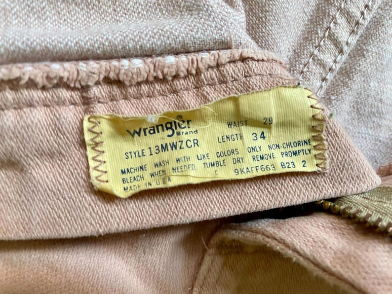 May include: A pair of light tan denim pants featuring a yellow interior tag with the Wrangler brand logo, style number 13MWZCR, and printed measurements of 29 inch waist and 34 inch length. The tag includes care instructions and Made in USA text.
